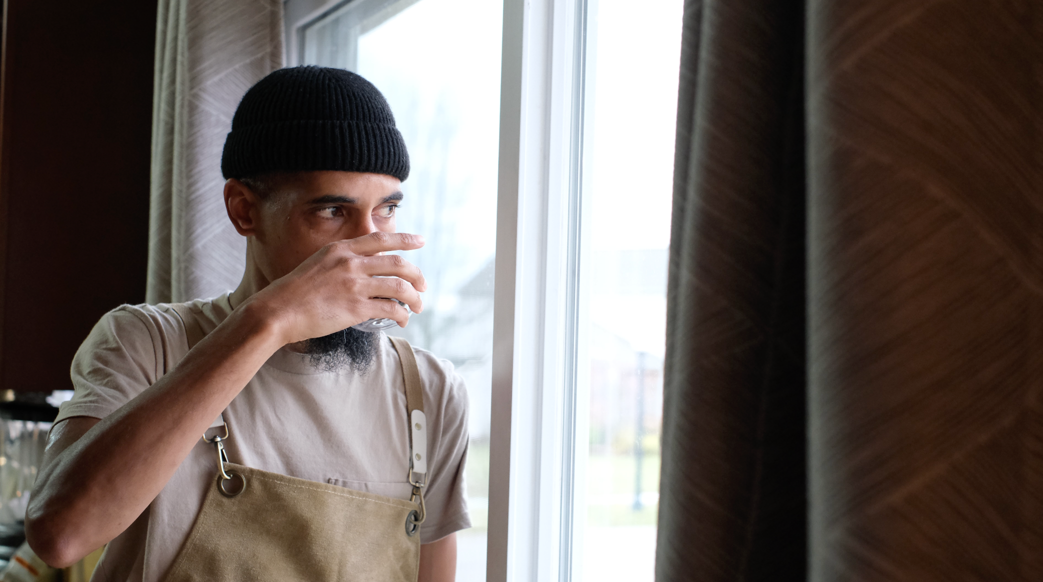 Founder, Michael Davis, wearing a brown apron and black beanie, sips coffee near a window with lots of sunlight.